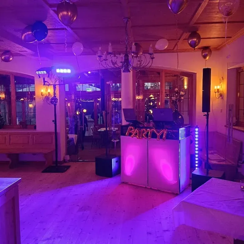 DJ-Pult mit goldenem Party-Schriftzug und Ballons im Gastraum, violett beleuchtet