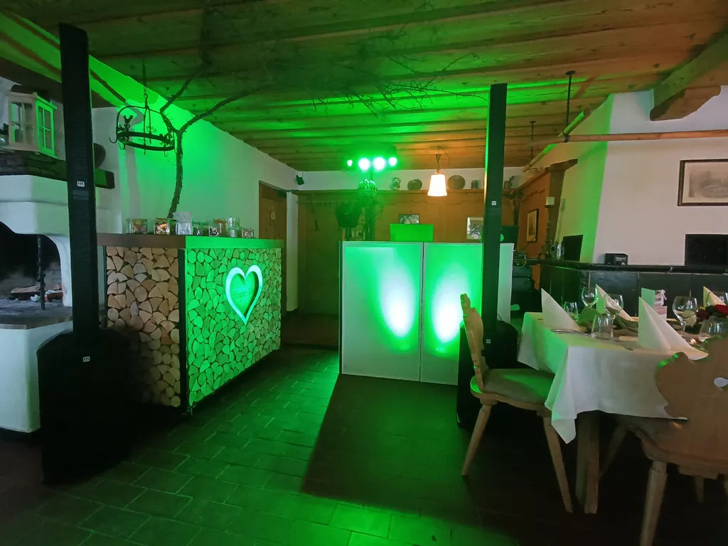 DJ-Rico-Cinsano DJ-Pult mit zwei Säulenlautsprechern und grünem Licht in der Gaststube der Stögeralm am Schliersee, daneben die gedeckte Hochzeitstafel
