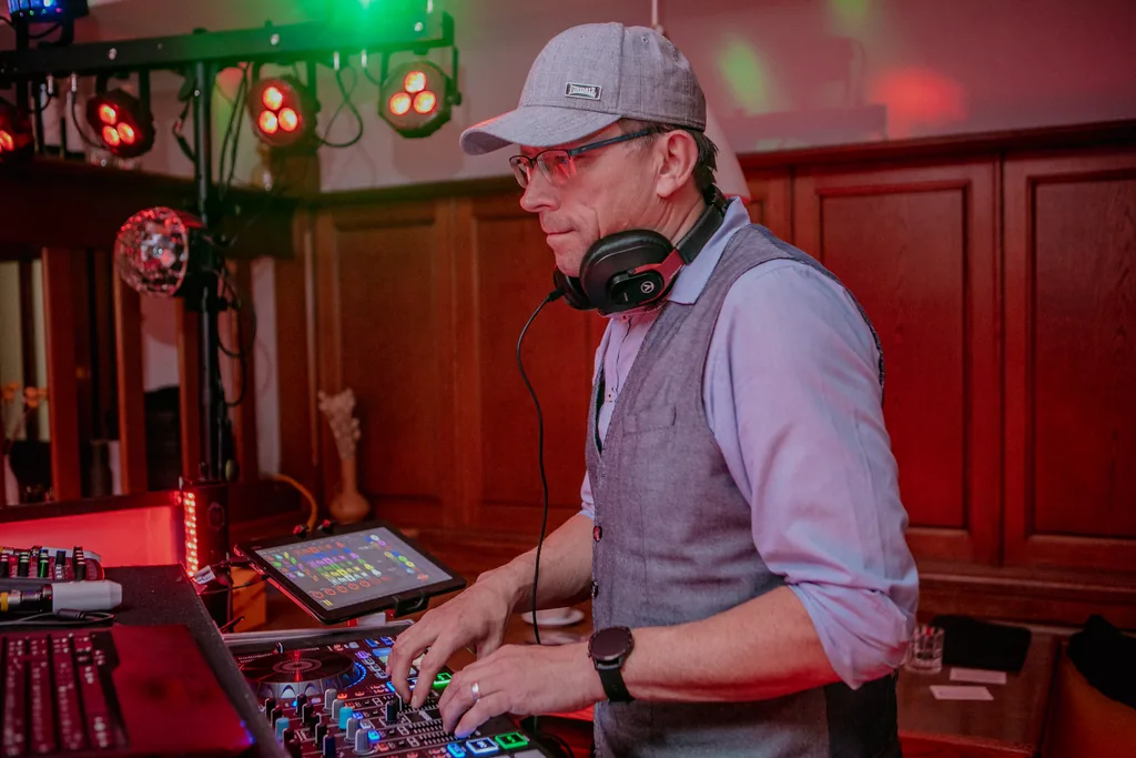 DJ Rico Cinsano mit grauer Cap und Kopfhörern am DJ-Controller, daneben ein Tablet für die Lichtsteuerung, dahinter Lichtstativ und Spiegelkugel im holzvertäfelten Saal