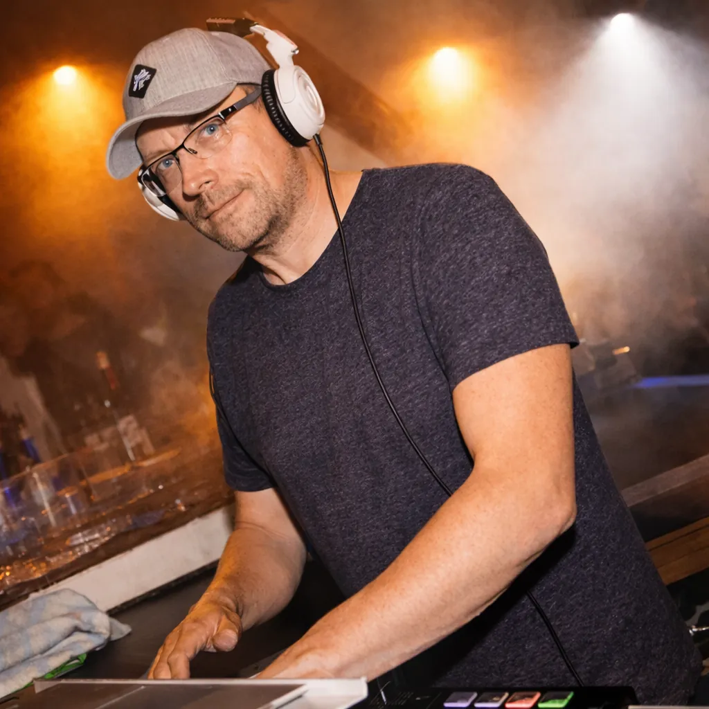 DJ Rico Cinsano mit Kopfhörern am DJ-Pult, dahinter Nebel und orange Scheinwerfer