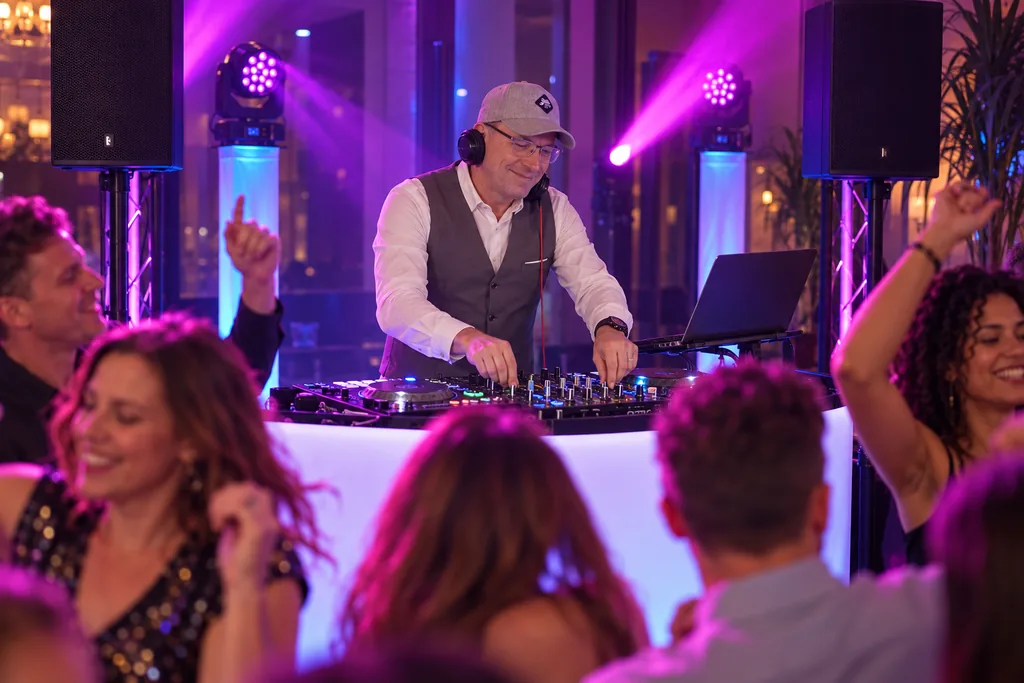 DJ Rico Cinsano in Weste am DJ-Pult zwischen zwei Lautsprechern und violetten Scheinwerfern, im Vordergrund tanzende Gäste