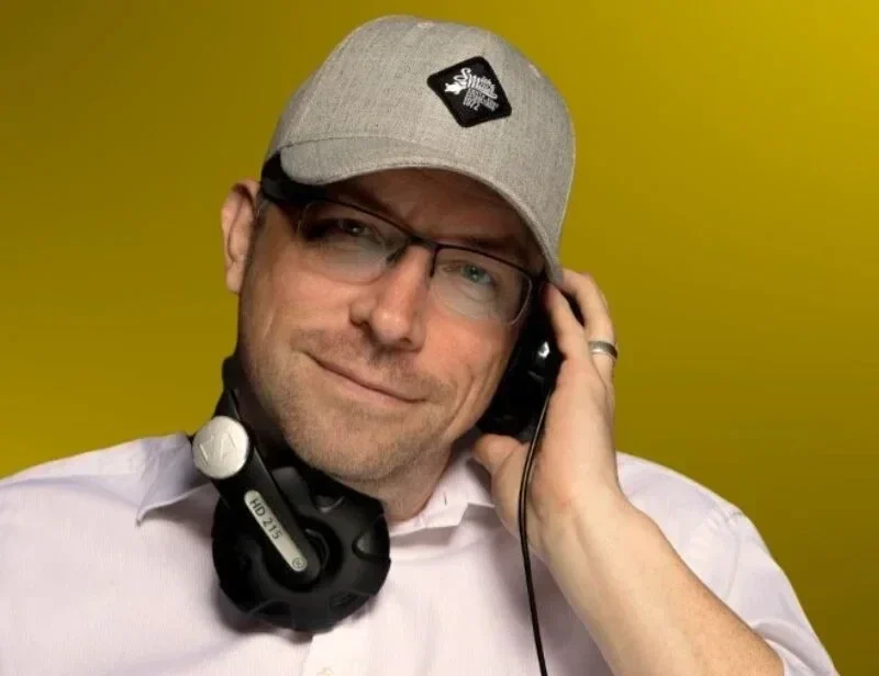 DJ Rico Cinsano mit grauer Cap, Brille und Kopfhörern vor gelbem Hintergrund