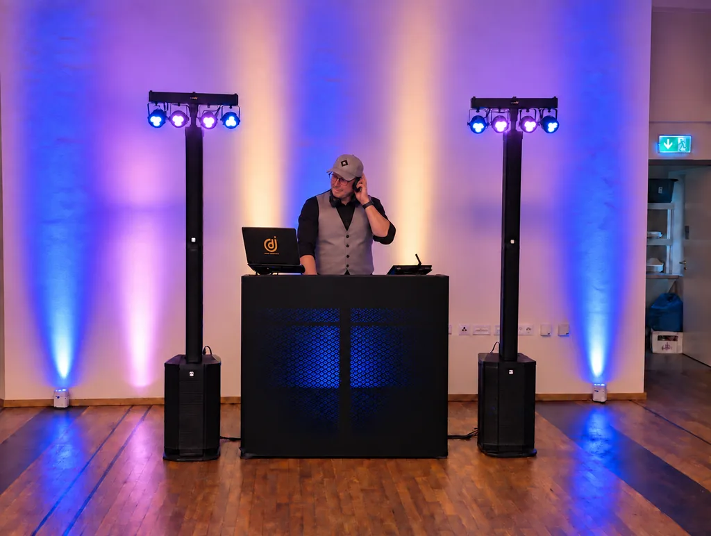 DJ Rico Cinsano mit Kappe und Kopfhörer am DJ-Pult, links und rechts Lichtstative mit Säulen-PA vor blau und orange angestrahlter Wand