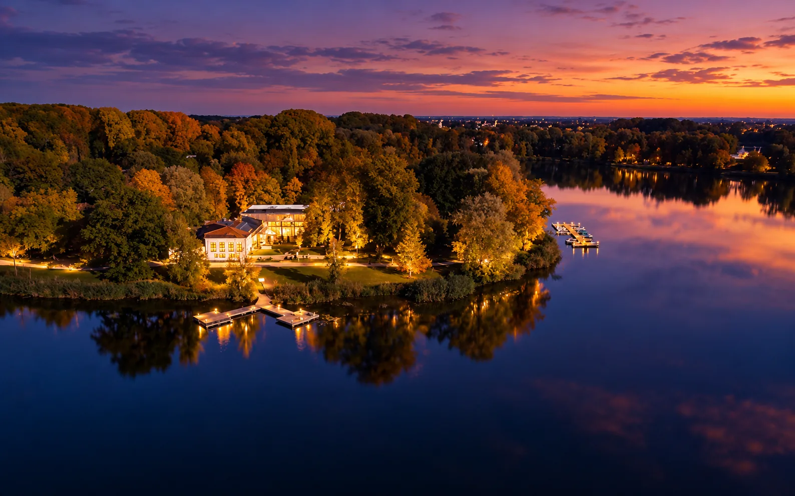 Seehaus Ingolstadt am Abend von oben: beleuchtetes Haus am Baggersee mit Steg, Herbstbäumen und Abendrot