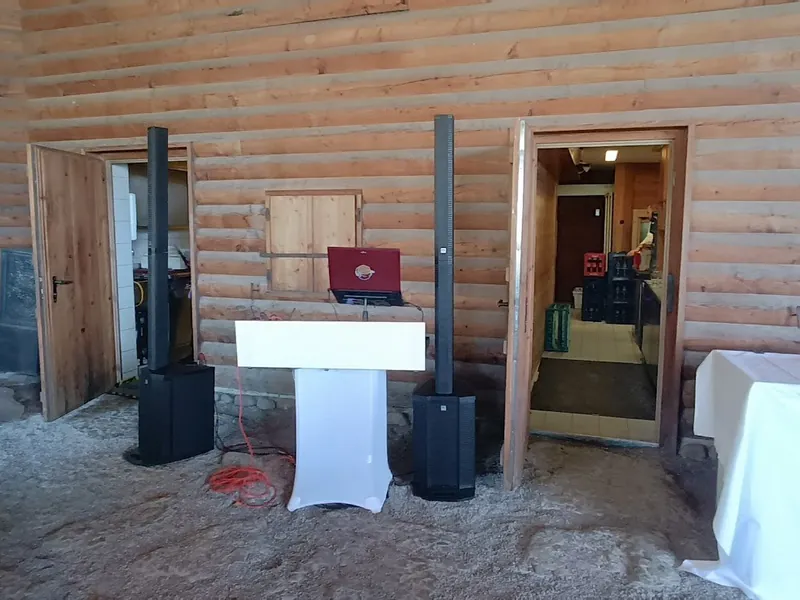 DJ-Setup mit Säulen-Lautsprechern und weißem DJ-Pult vor Holzwand der Sonnenkopfhütte